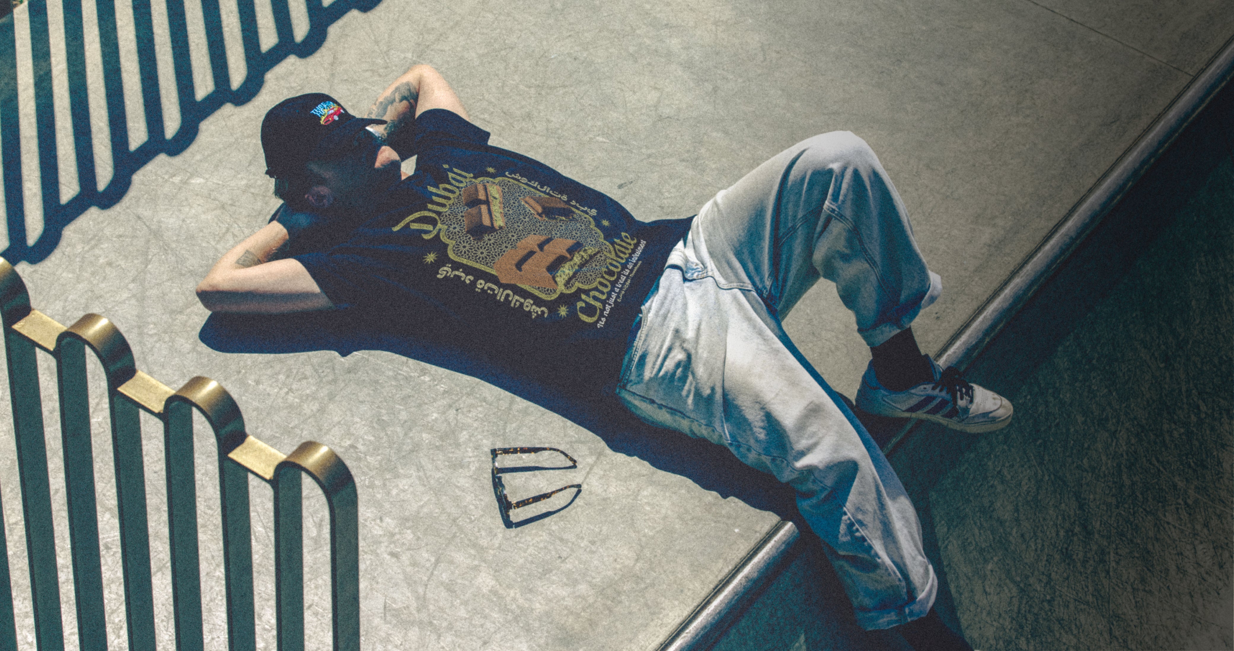 Person lying on a concrete surface wearing a black t-shirt with a graphic design and white pants.
